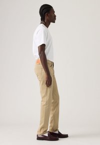 Μπεζ βαμβακερό παντελόνι με ίσια κοπή, πλαϊνές τσέπες και πλακέ ετικέτα στη πίσω ζώνη, φορεμένο με λευκό μπλουζάκι και καφέ μοκασίνια.