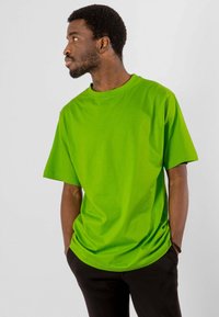 Mand med skæg og afrofrisure, iført en lys grøn t-shirt og sorte bukser, står med hænderne i lommerne og ser til venstre.
