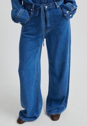 Blå højtaljede jeans med vide ben og bindesnøre i taljen, kombineret med en matchende denimjakke og brune sko med firkantet tå.