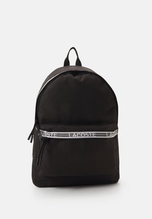 Mochila de tela negra con forma redondeada, que presenta una banda con el logo blanco de Lacoste, doble cremallera y un asa en la parte superior para llevar.