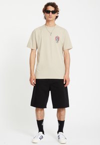 Beige cotton t-shirt featuring a logo graphic, paired with black denim shorts. Model wears dark sunglasses and white sneakers.