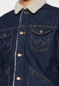 Džínová bunda se světle béžovým límcem z umělé beránkové textilie, měděným kováním, dvěma náprsními kapsami a kontrastním šitím v tmavě modré barvě.