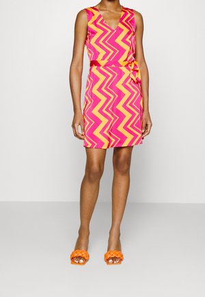 Levendige mouwloze jurk met een zigzagpatroon in roze en geel, voorzien van een V-hals en een ceintuur in de taille. Gecombineerd met oranje gevlochten instapschoenen.