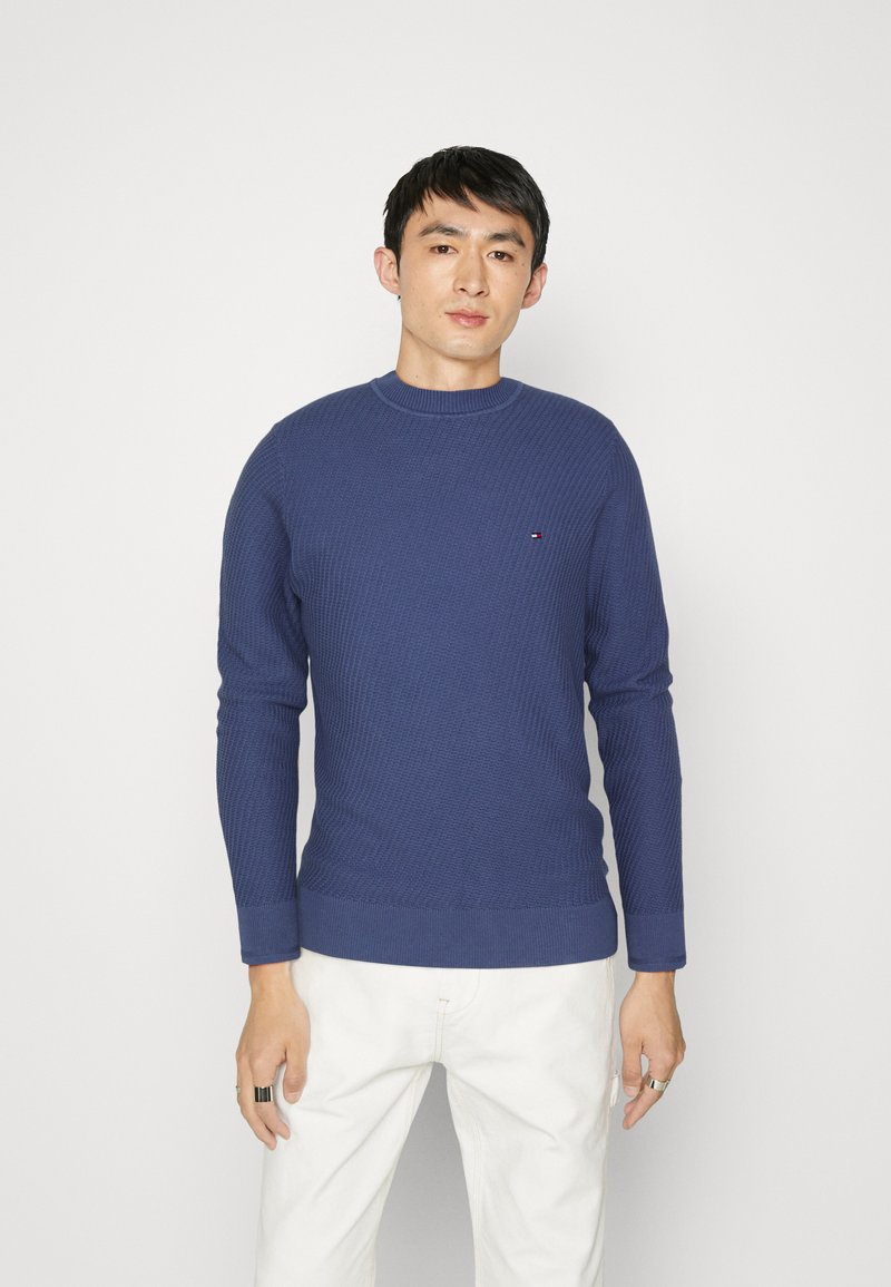 Maglione a costine blu navy con colletto a girocollo e polsini aderenti, con un piccolo logo sul petto, abbinato a pantaloni chiari.