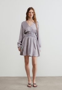 Γυναίκα με μωβ ανοιχτό μανίκι φόρεμα με ζώνη στη μέση, που κρατάει ασημένιο τσαντάκι, στέκεται σε απλό πάτωμα μπροστά σε λευκό τοίχο.