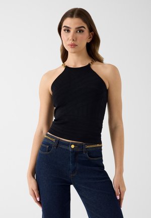 Jeune femme portant un haut noir sans manches avec des bretelles en chaîne dorée et un jean bleu foncé avec un détail de ceinture chaîne dorée.