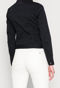 Black denim jacket with a fitted design, button accents on the cuffs, and a cropped length; paired with white jeans showing back pockets.