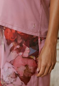 Ružová blúzka s kvetinovým vzorom na spodnej časti. Hladká textúra, voľný strih, obsahuje malé zlaté logo na leme. Elegantný dizajn.