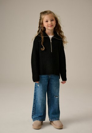 Schwarzer gestrickter Pullover mit einem halben Reißverschlusskragen, kombiniert mit hellblauen, ausgewaschenen weit geschnittenen Jeans und beigen Slippers.