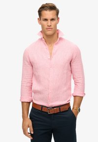 Camisa de botones a rayas rosas con el collar levantado, mangas largas remangadas en los puños, combinada con un cinturón marrón y pantalones oscuros.