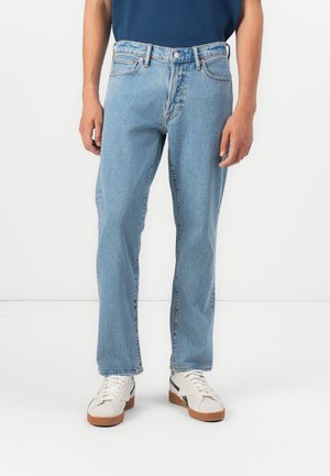 Light blue denim jeans with a straight cut, subtle texture, five pockets, and stitched accents. Worn with white sneakers with brown soles.