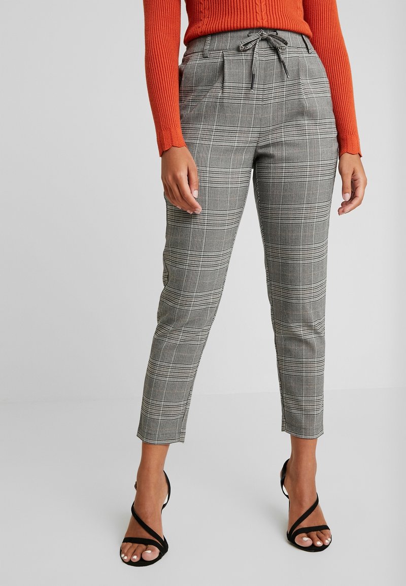 Pantaloni a scacchi grigi con un design aderente, vita alta, tasche laterali e chiusura con cordoncino. Indossati con tacchi neri a fascia.