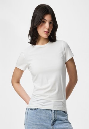 Femme aux cheveux foncés mi-longs, portant un t-shirt blanc uni à manches courtes et un jean bleu clair, debout les mains dans le dos.