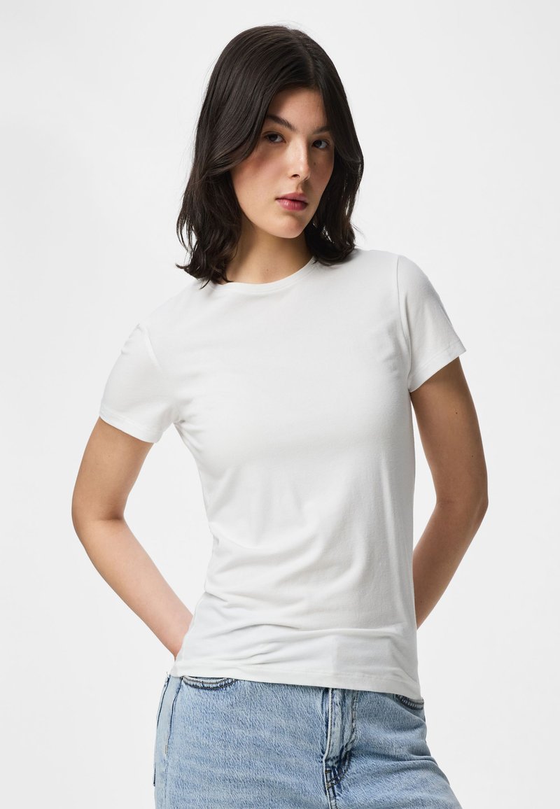 Femme aux cheveux foncés mi-longs, portant un t-shirt blanc uni à manches courtes et un jean bleu clair, debout les mains dans le dos.