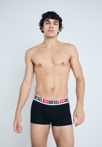 Boxer-briefs noirs avec une ceinture blanche portant "DIESEL" en lettres rouges. Modèle avec des cheveux bouclés se tenant contre un fond uni.
