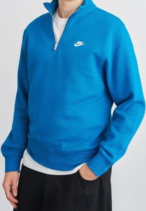 Mann trägt ein hellblaues Nike-Quarter-Zip-Sweatshirt über einem weißen Hemd mit schwarzer Hose, eine Hand in der Tasche und die andere entspannt.