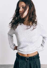 Žena s vlnitými hnedými vlasmi nosí svetlosivý krátky sveter a tmavé nohavice s vysokým pásom a čiernym opaskom, stojaca pred jednoduchou bielou stenou.