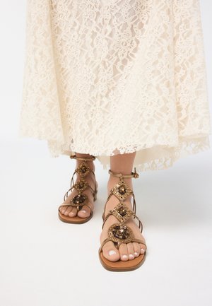 Feet wearing tan gladiator sandals with decorative gold and brown jewels, under a cream lace dress hem.