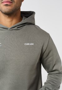 Άνδρας με γκρι φούτερ με κουκούλα, με το κείμενο "CLSR LDN" τυπωμένο με λευκά γράμματα στο στήθος, εμφανιζόμενος από τον λαιμό έως το μέσο του κορμού.
