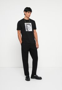 Black cotton t-shirt featuring a graphic print within a white rectangular border, paired with black trousers and black shoes.