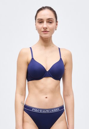 Młoda kobieta w granatowym biustonoszu i dopasowanej bieliźnie Polo Ralph Lauren, stojąca na jednolitym, jasnoszarym tle.