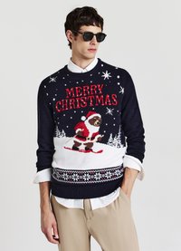 Maglione di Natale blu navy con un orso che scia vestito da Babbo Natale, con la scritta "BUON NATALE" decorata con motivi di fiocchi di neve rossi e bianchi.