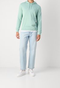 Mátově zelená mikina s kapucí na stažení, přední kapsou a měkkou texturou; kombinovaná s světle modrými džíny s rovnými nohavicemi a bílými teniskami.