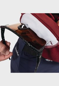 Sonnenbrille mit einem schwarz gesprenkelten Rahmen und braunen Gläsern, gehalten in der Nähe eines kastanienfarbenen Fleece, das das geschwungene Design und die integrierte Hardware zur Geltung bringt.