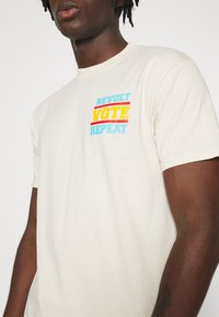 Béžové bavlněné tričko s barevným potiskem: "REVOLT VOTE REPEAT" v tyrkysové, žluté a červené barvě. Krátké rukávy, klasický výstřih.