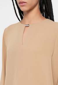 Bluză bej cu un decolteu subtil în V și un accent metalic la guler. Țesătura este fină, cu un ușor luciu, având mâneci lungi.