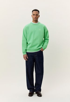 Ung mand stående iført en lys grøn sweatshirt, mørkeblå jeans og mørkebrune sko mod en ensfarvet lys baggrund.