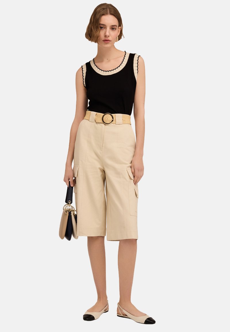 Czarna bezrękawnikowa bluzka z falbaniastym dekoltem, beżowe szorty cargo, szeroki pasek i jasne buty z czarnymi noskami. Modelka trzyma torebkę.