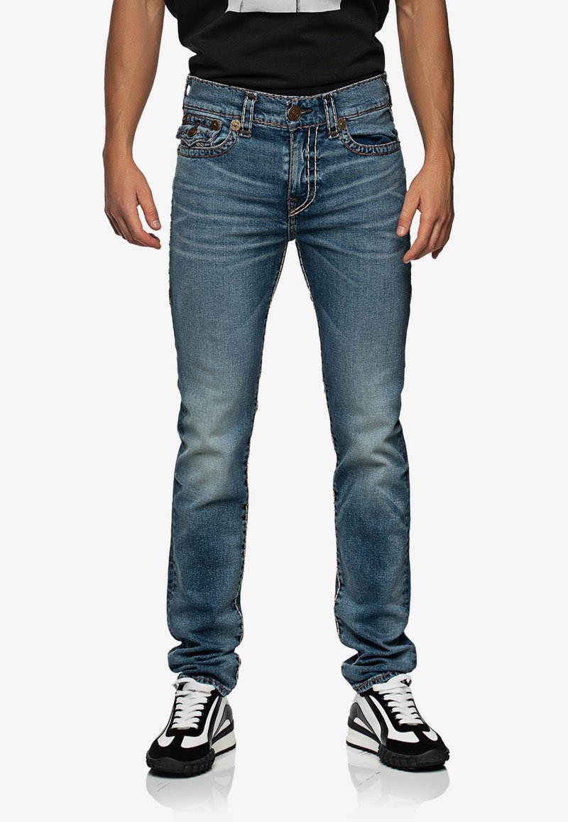 Homme portant un jean bleu délavé, des baskets noires et blanches, et une chemise noire, debout, mains détendues le long du corps sur fond blanc.