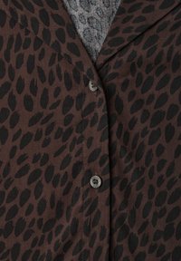 Brown button-up shirt with black irregular spots worn over a gray patterned fabric visible at the collar.