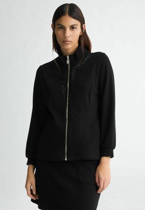 Veste zippée noire avec un col montant, ornée d'accents ressemblant à des perles, et poches avant. Texture douce et silhouette ajustée.