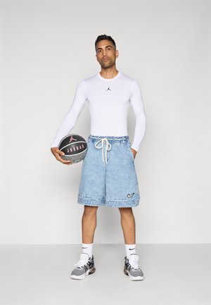 Man in white long-sleeve shirt and light denim shorts holds black and gray Jordan basketball, standing against plain light background.
