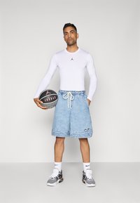 Mies, który ma na sobie białą, długą koszulę z długim rękawem oraz jasne dżinsowe szorty, trzyma czarno-szary kosz do koszykówki Jordan, stojąc na tle jednolitego, jasnego tła.