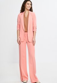 Różowy skrojony garnitur z długim żakietem i spodniami o szerokich nogawkach. Materiał ma gładką teksturę, a spodnie wyposażone są w podwójne zapięcie na guziki.