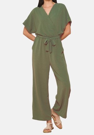Frau trägt einen olivgrünen Kurzarm-Jumpsuit mit gebundener Taille und weitem Bein, kombiniert mit beigen Sandalen mit offenen Zehen.