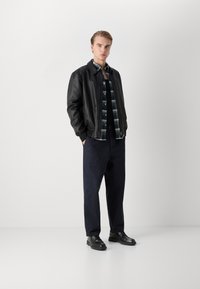 Zwarte leren jas over een geruite shirt, gecombineerd met donkere broek en zwarte schoenen. Model staat op een witte achtergrond.