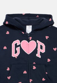 Tumši zila jaundzimušo kapuce ar rozā sirds rakstu un lielajiem burtiem "G" un "P", ko krustojas rozā sirds uz krūtīm, priekšējās rāvējslēdzējs.