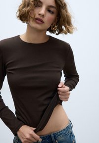 Maglia marrone a maniche lunghe in materiale liscio, design aderente, scollo rotondo, abbinata a jeans di denim blu. Pelle visibile all'altezza del ventre.