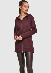 Bordo zip-up haljina s kapuljačom, zlatnim patentnim zatvaračem, prednjim džepovima i dugim rukavima. Mekani materijal i prilagođen dizajn s dužinom do sredine butina.