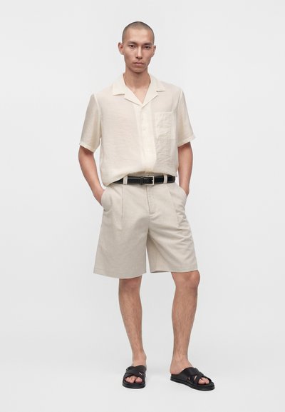 Homme portant une chemise beige à manches courtes boutonnée, un short assorti avec une ceinture noire, et des sandales noires, debout les mains dans les poches.