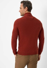 Roter, strukturierter Pullover mit langen Ärmeln und einem umgeschlagenen Kragen. Verfügt über einen gerippten Saum und ein gesprenkeltes Muster, getragen über beigefarbenen Cordhosen.