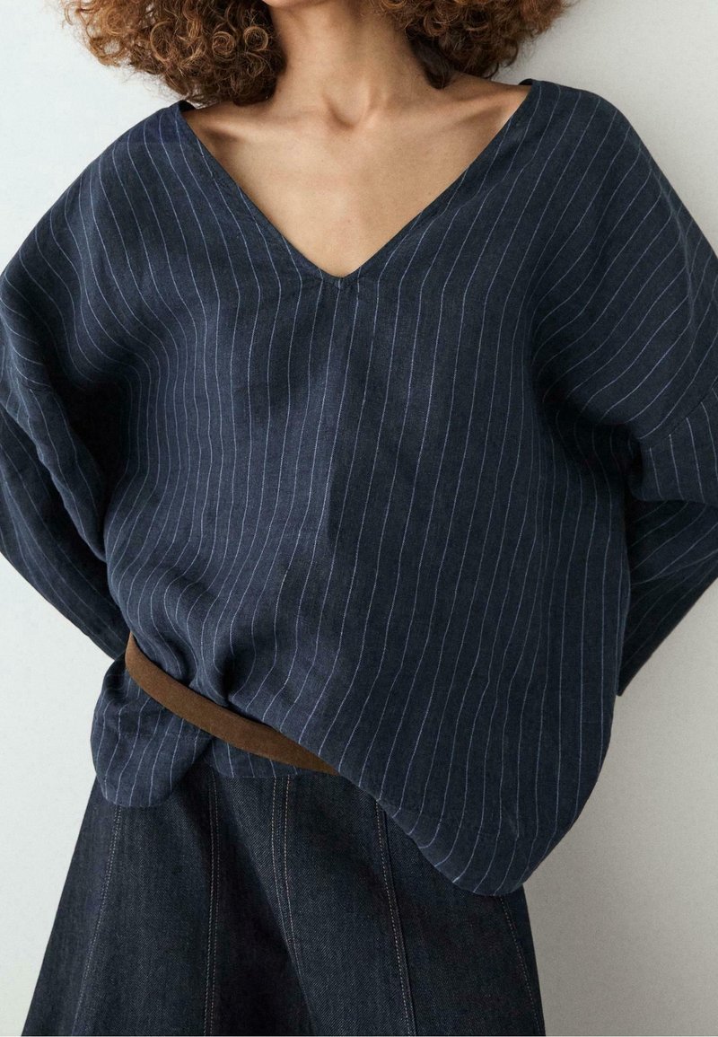 Personne aux cheveux bouclés portant un haut bleu marine à fines rayures avec un col en V, rentré dans un pantalon foncé taille haute avec une ceinture marron.