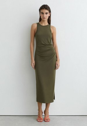 Žena stojaca pri bielej stene, oblečená v olivovo zelenej midi šatke bez rukávov s bočným rozparkom a koralovými otvorenými lodičkami.