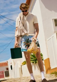 Polo beige clair texturé, short de bain bleu à motif tropical, tenant un sac en filet avec des citrons, pieds nus avec des chaussettes blanches et des mocassins marron.