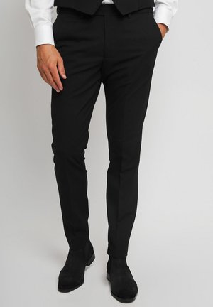 Mann trägt schlanke schwarze Anzughose, schwarze Schuhe, weißes Hemd und schwarze Weste, steht mit einer Hand in der Tasche.