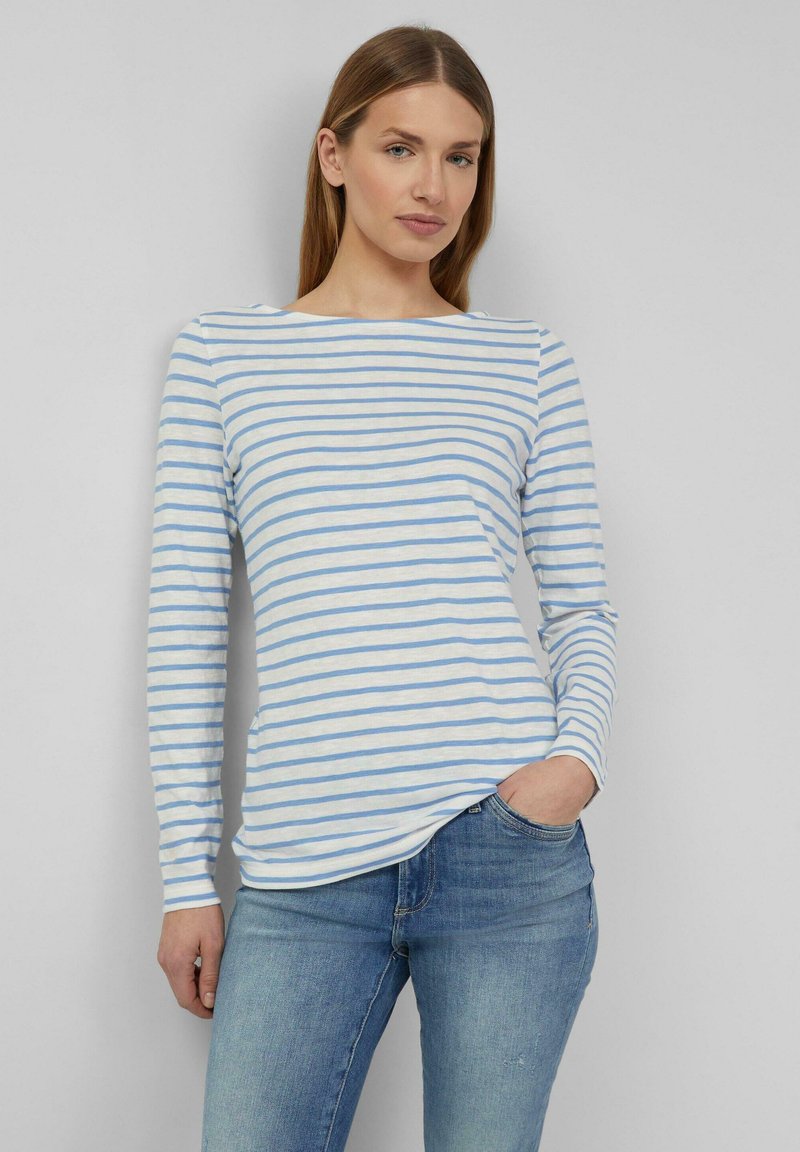 Moteris vilki ilgomis rankovėmis baltą marškinį su šviesiai mėlynomis horizontaliomis juostomis ir mėlynas džinsas, stovi viena ranka kišenėje.